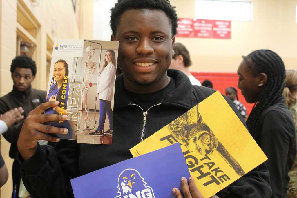 Man in a hallway, holding a book, smiling. In background, two people holding a yellow and a blue book.