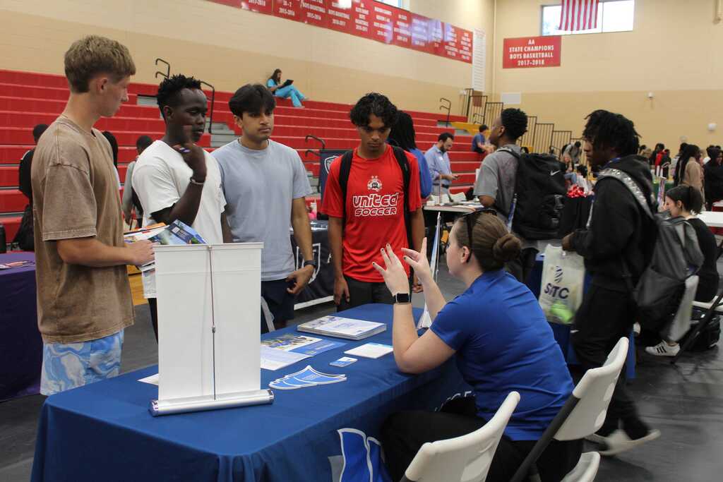 Several young adults stand in line at a table with a woman in a blue shirt.