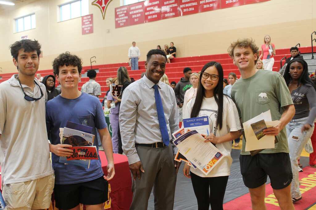 Five people stand together in a school gym. Each holds a book. They smile, standing before a blurred background.