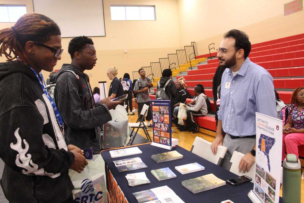 People at a table with pamphlets and items, one person looking at a man in a blue shirt.