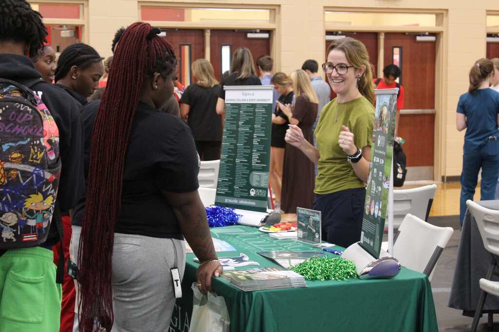 A woman with glasses stands by a table with a green cover; others gather nearby. In background, people mill around in a corridor.