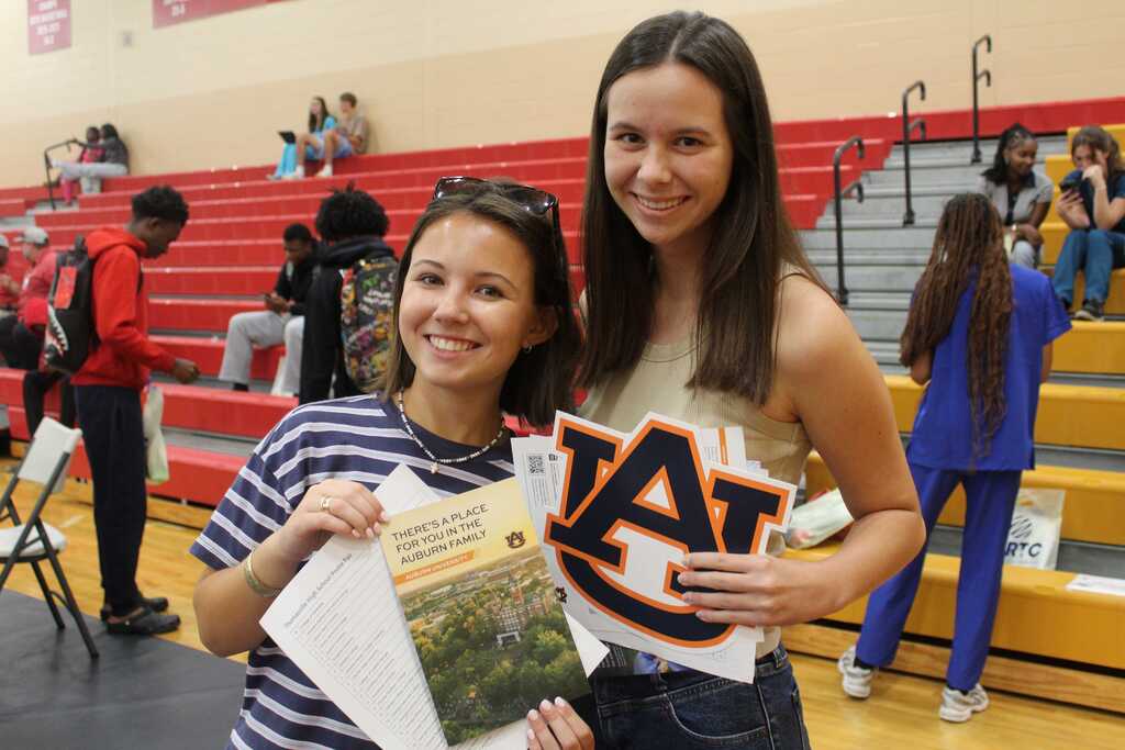 Two women stand in front of bleachers, one holding papers, the other wearing a white and blue top.