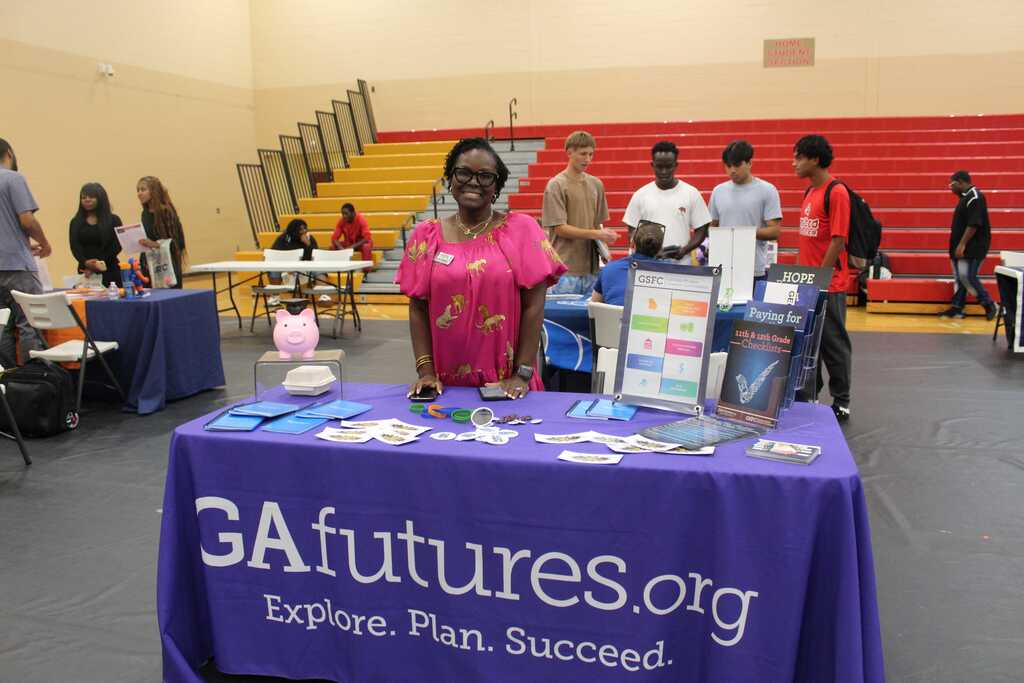 A person stands at a table with a purple cloth. The tablecloth reads "GAfutures.org Explore Plan Succeed". People are present in the background.