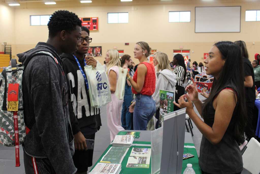 Several people are standing and talking at an indoor event. A woman speaks with two men. A green table holds brochures.