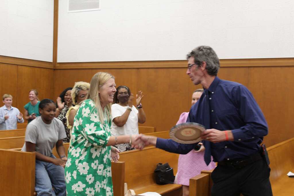Two people exchange a handshake and an object in a room with wooden benches. Several people are seated and standing behind them.