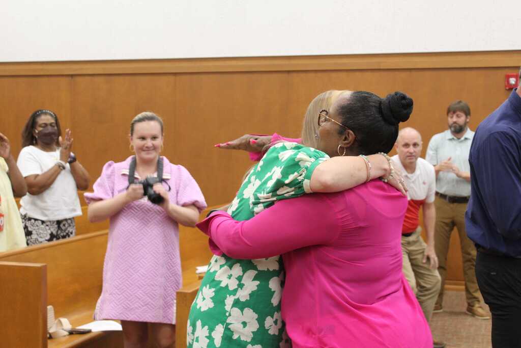 Two women in green and pink dresses embrace each other while several people observe in a room with wooden paneling.