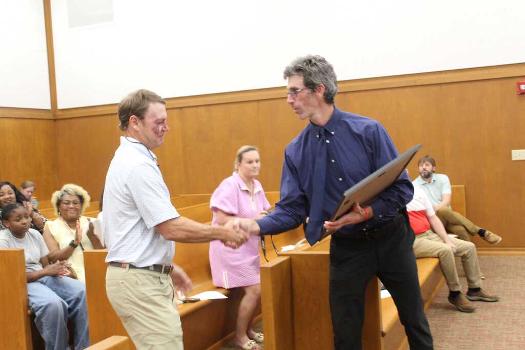 Two men shake hands with each other; one holds an award. Behind them, several people sit in wooden benches.