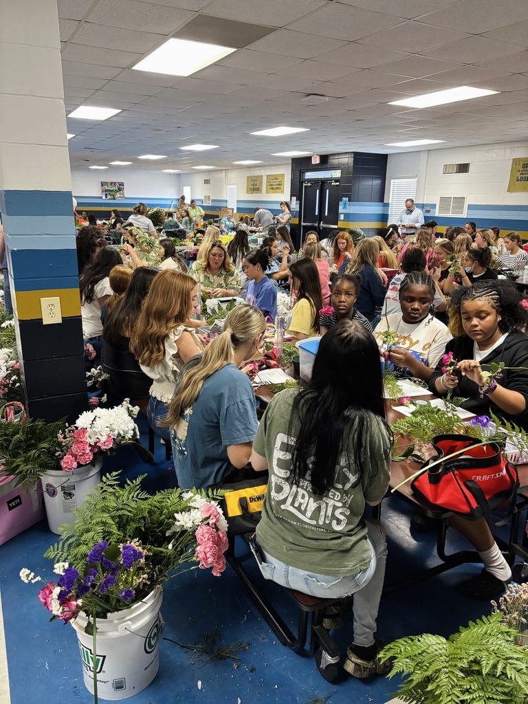 A large group of people seated around tables, arranging flowers in pots, inside a room.