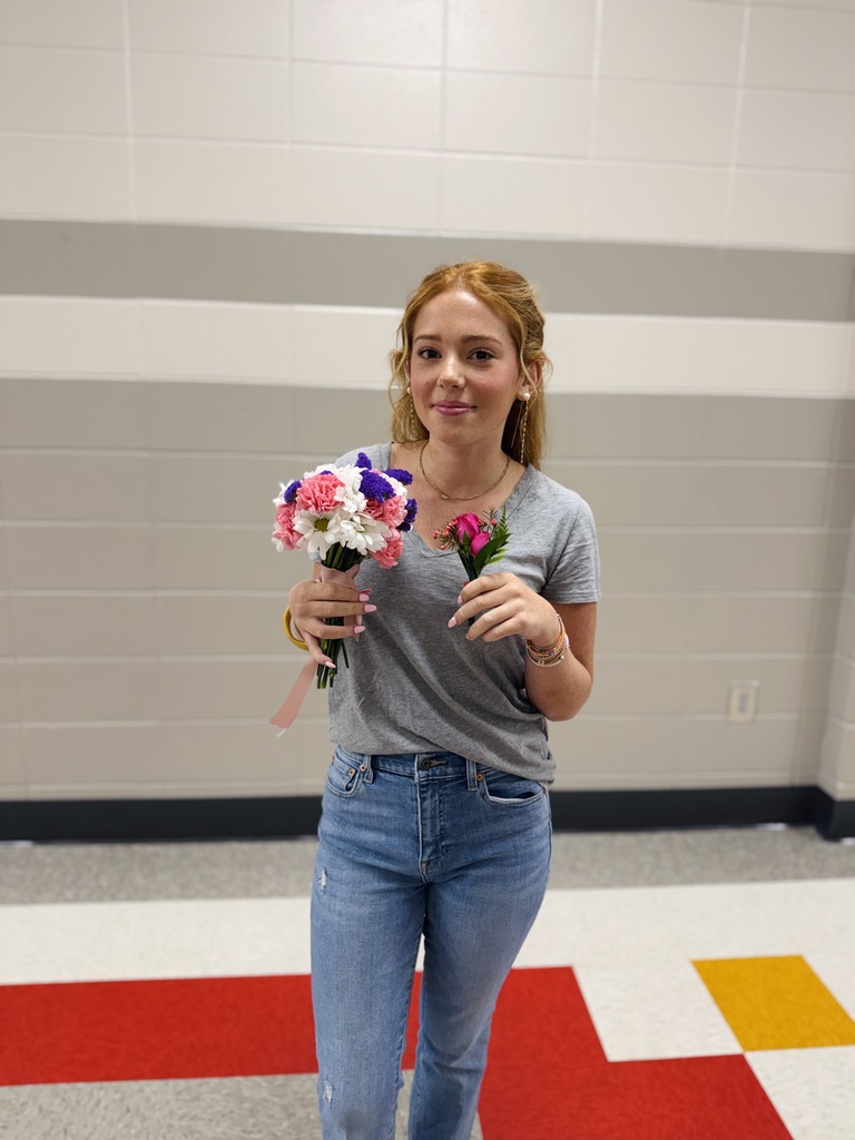 A woman holding a bouquet and a single flower stands in a hallway with tiled floors and white walls.