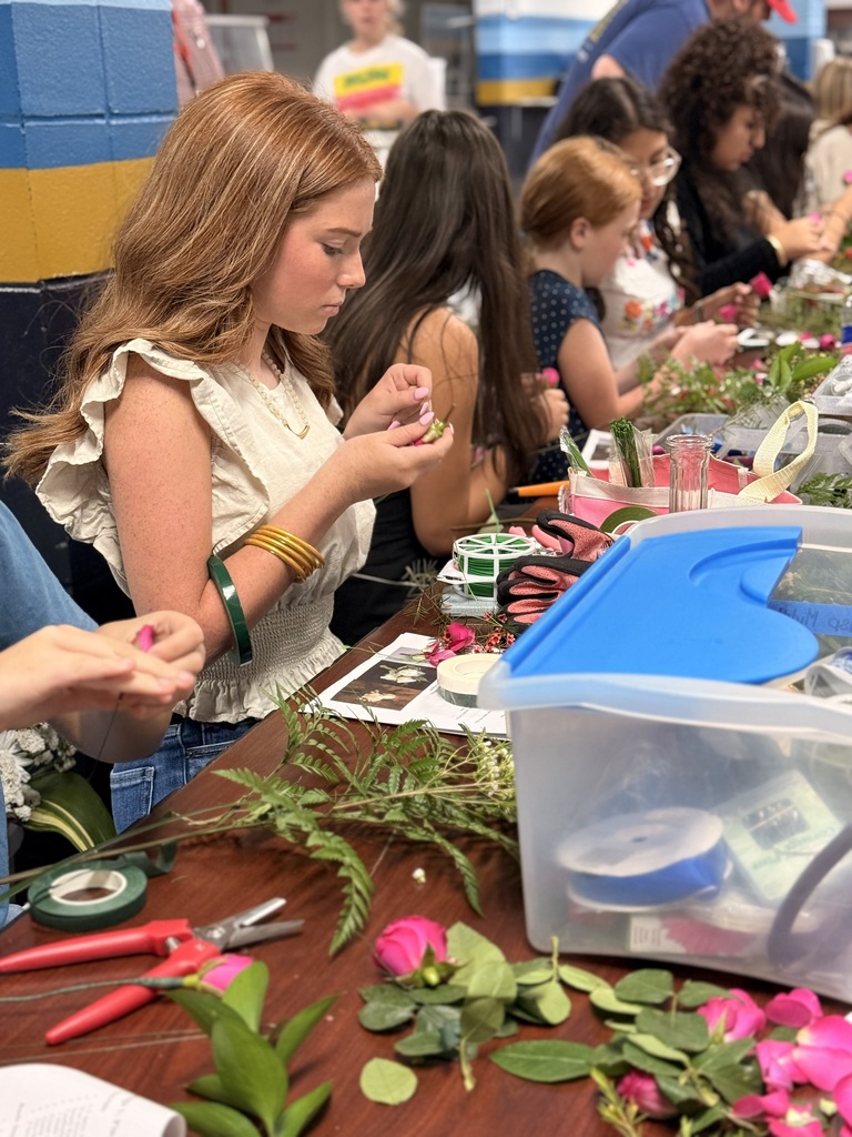 People seated at a table, arranging flowers, leaves, and ribbons. A clear container with tools and a green bracelet.