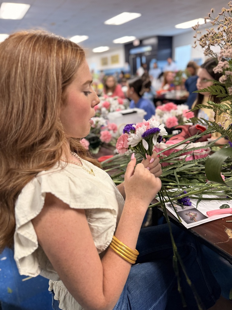 A woman with long hair in a white blouse, seated, carefully arranging flowers. A table filled with flowers is in front. Background shows other people working.