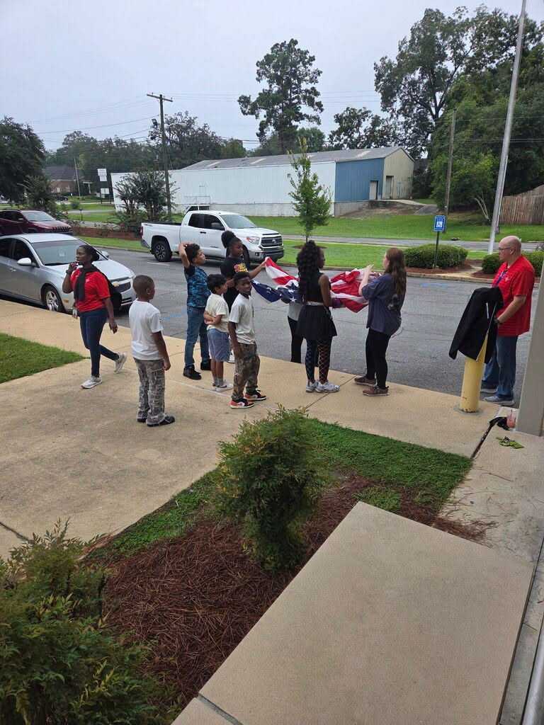 People stand on a sidewalk, holding an American flag. Some wear red, and one wears a backpack. Cars and a pickup truck are parked. Trees and bushes are in the background.