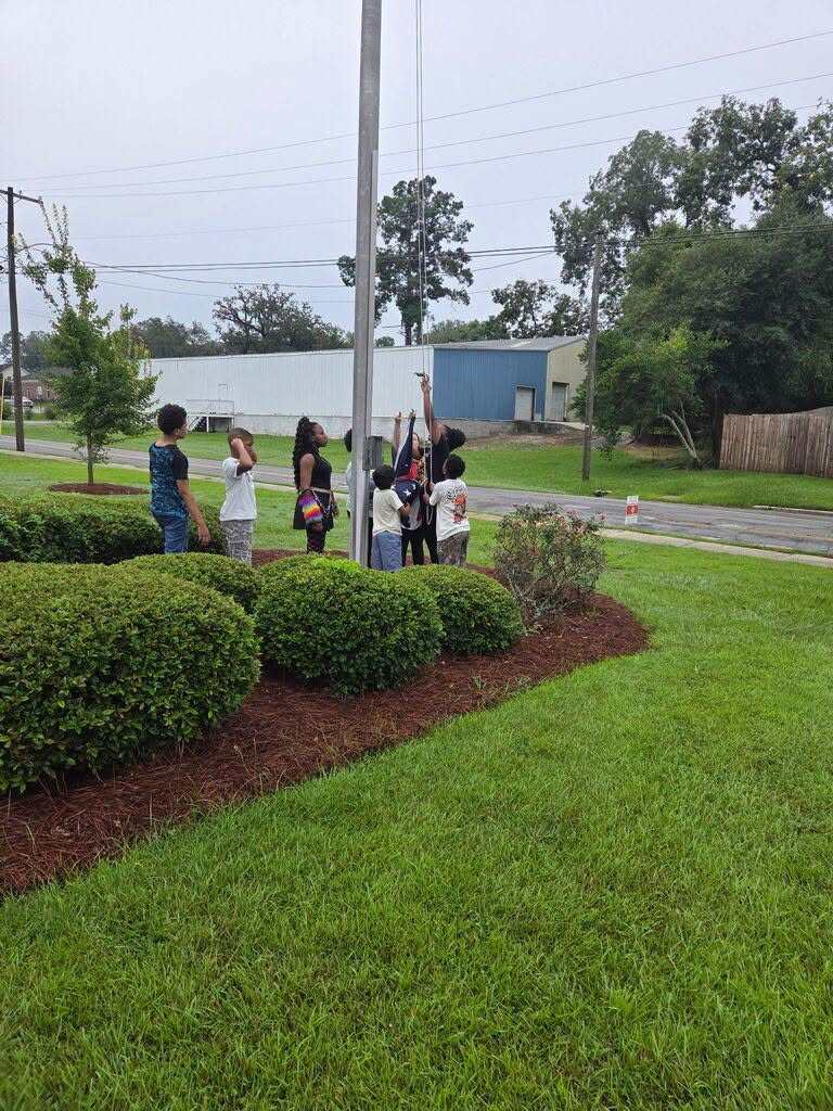Group of people standing around a flagpole in a grassy area with bushes and trees. A building is visible in the background.