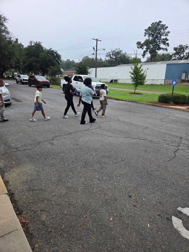 People walk across a road with cars passing by. There is a white arrow on the pavement.
