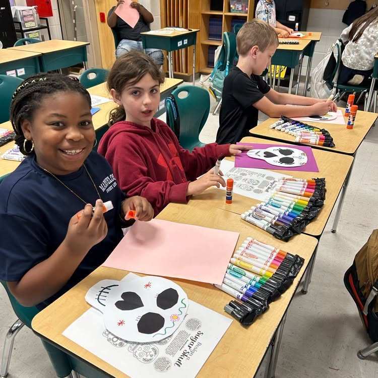 Students sit at desks in a classroom, smiling, with art supplies and papers in front of them.