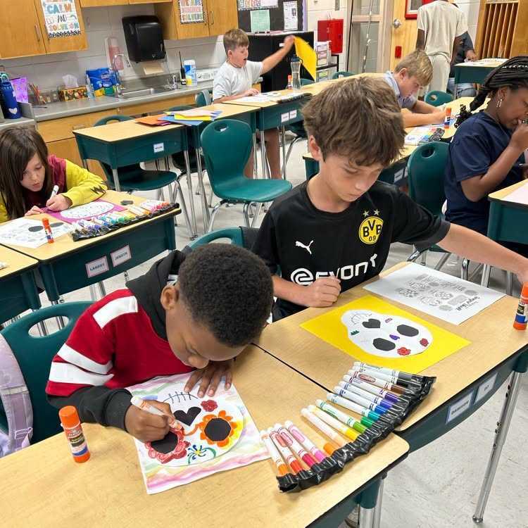 Kids in a classroom. They are sitting at desks and coloring a page. A boy has a completed coloring page in front of him.
