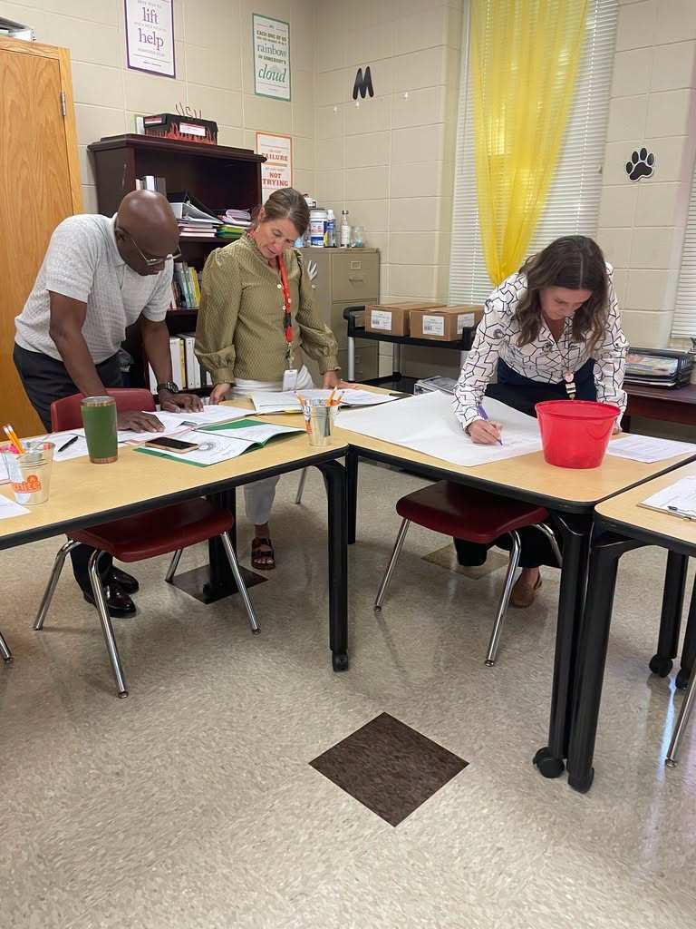 Three people in a classroom with tables. One bends over, another stands and writes, the third sits and writes. Papers and a bucket are on the tables.