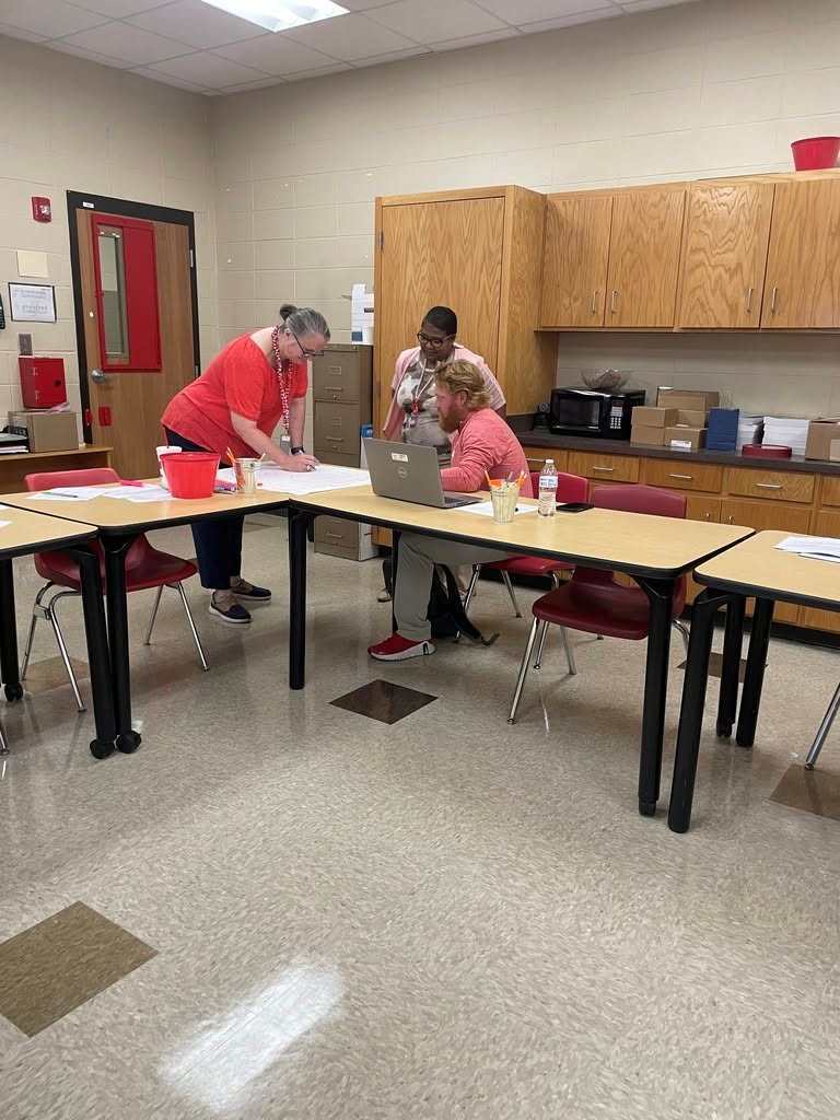 Three people in a classroom with desks, reviewing papers and using a laptop. Behind them are wooden cabinets.