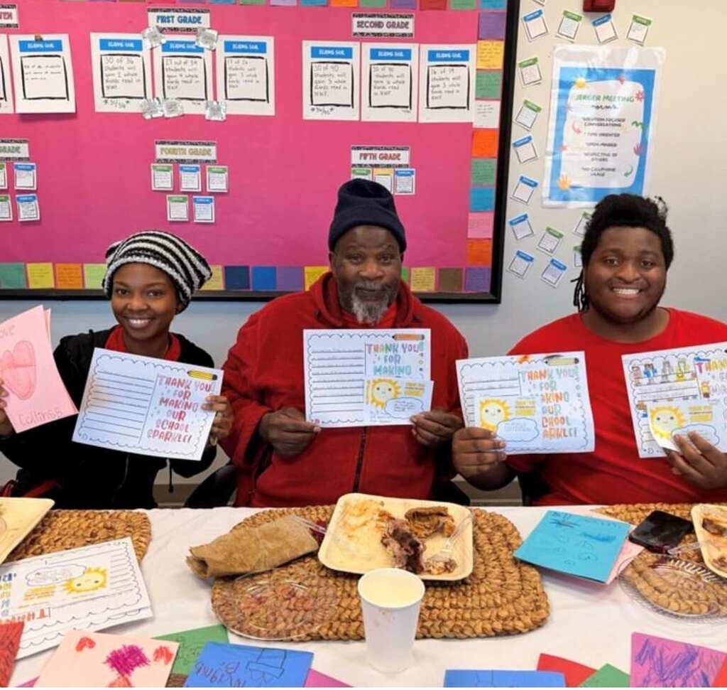 Three people pose with papers, smiling, in front of a colorful bulletin board with food on a table.