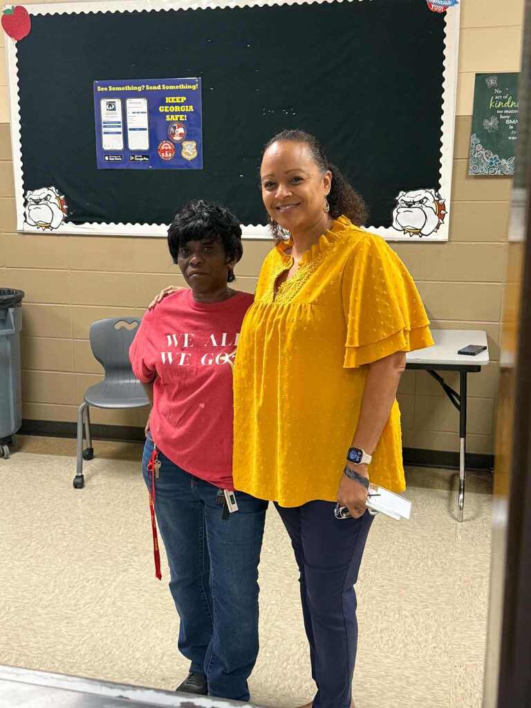 Two women stand side by side in a classroom. One wears a red shirt and blue jeans, and the other wears a yellow shirt.