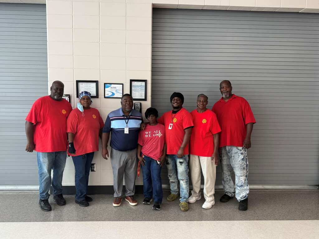 A group of seven people in red shirts pose in front of a garage with gray doors.