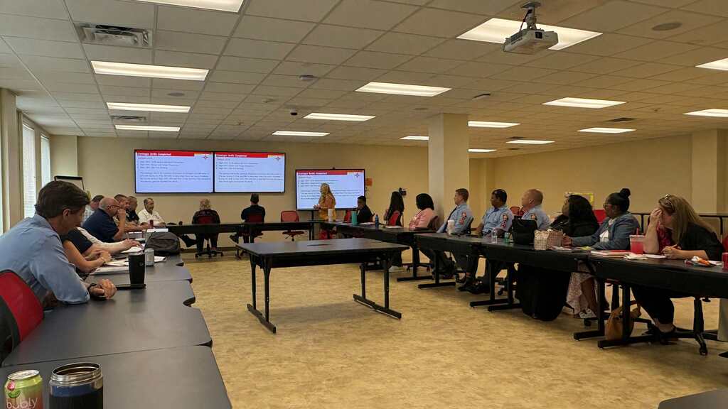 A group of people sitting in rows at a conference with tables and screens on a stage.