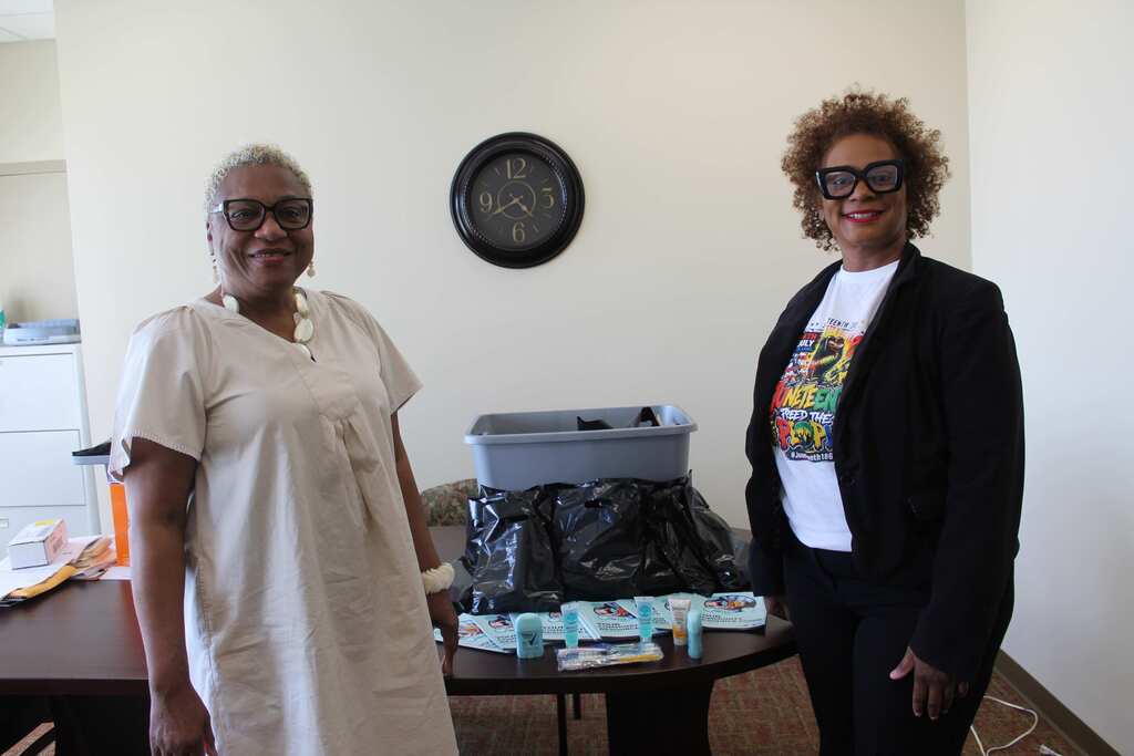 Two women stand near a table in an office. A clock is on the wall. One wears glasses and a white shirt, the other a black jacket and glasses.