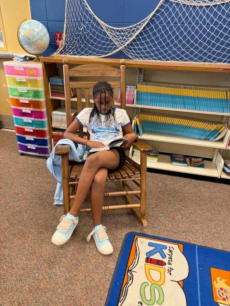 A young girl sits in a rocking chair, reading a book. She wears a white t-shirt, shorts, and white shoes.