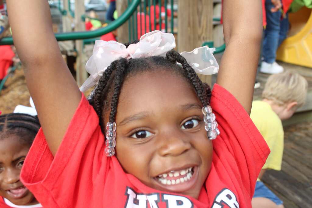 A smiling girl in a red shirt, hands raised, stands with a friend on a playground with a yellow slide.