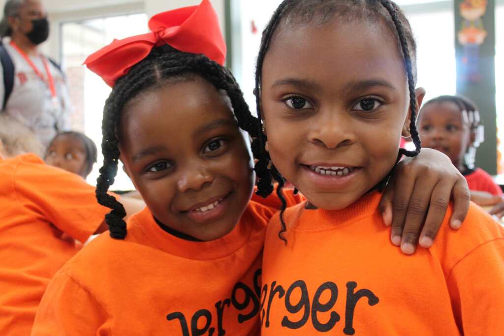 Two girls in orange shirts pose for a photo. One girl has a red bow in her hair.