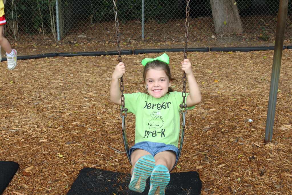 A young child wearing a green shirt sits on a swing, smiling, with a tree and fence in the background.