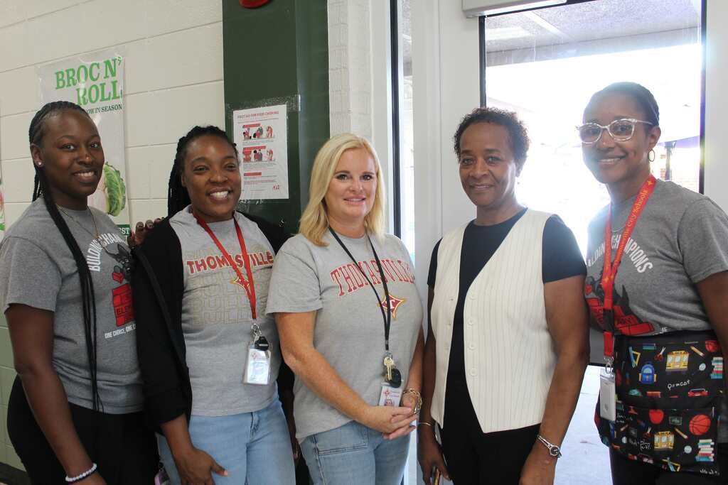 Five people stand in a room, wearing matching gray T-shirts. The woman in the center has a lanyard around her neck.