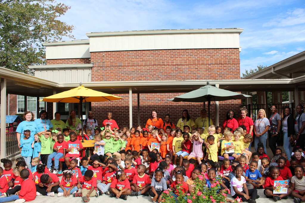 Large group of children and adults in front of a brick building with trees. Some wear red shirts.