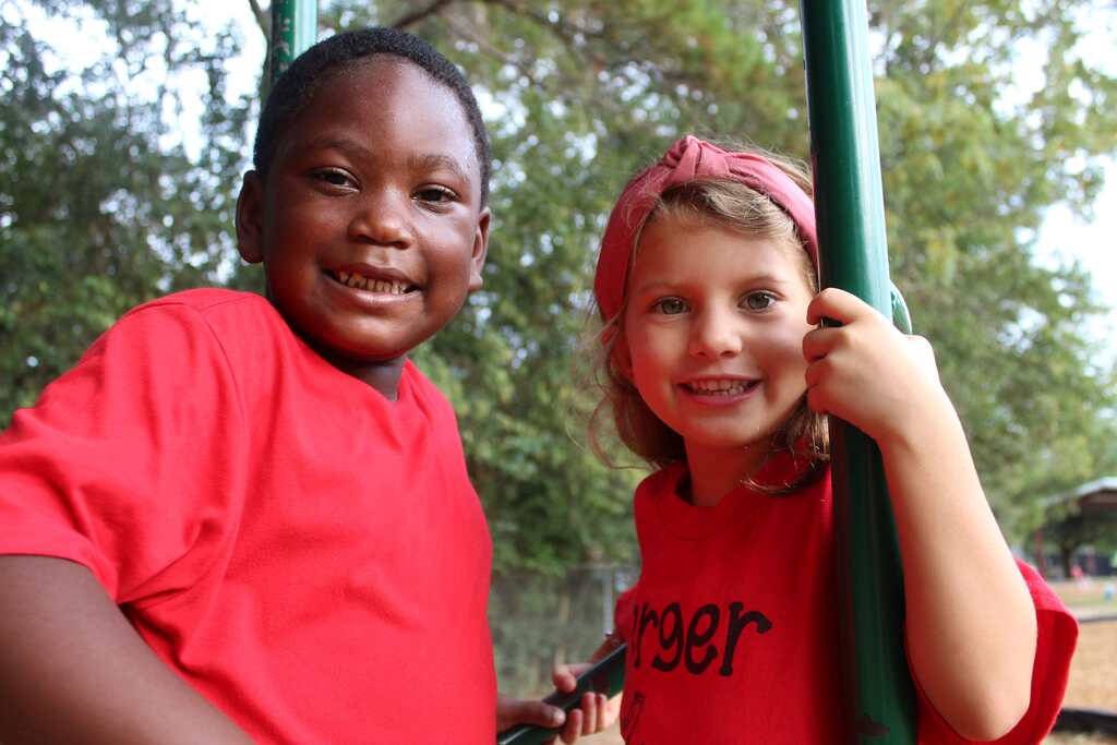 Two children in red shirts smile in a park. One holds a green swing. Trees and a fence are in the background.