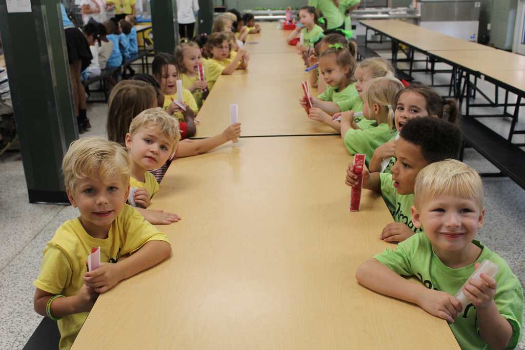 Children in matching yellow shirts seated in rows at a long cafeteria table. Some are holding utensils.