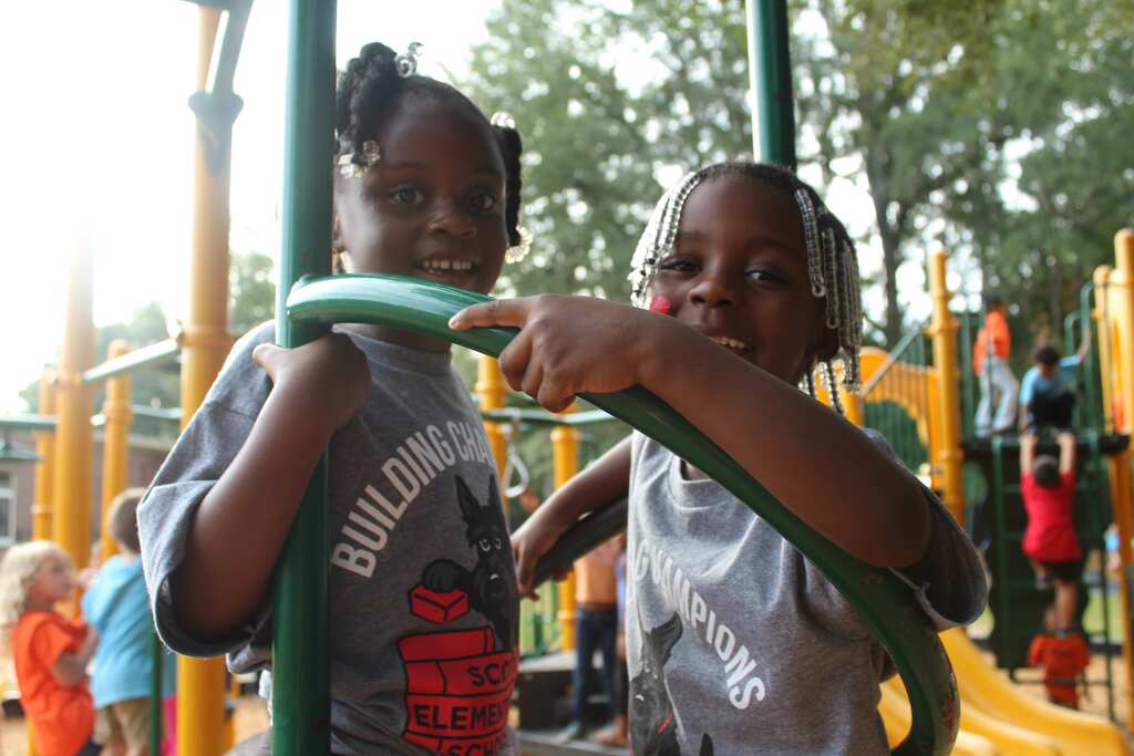Two children at a playground. One holds a green bar while the other points. Various playground equipment and trees in the background.