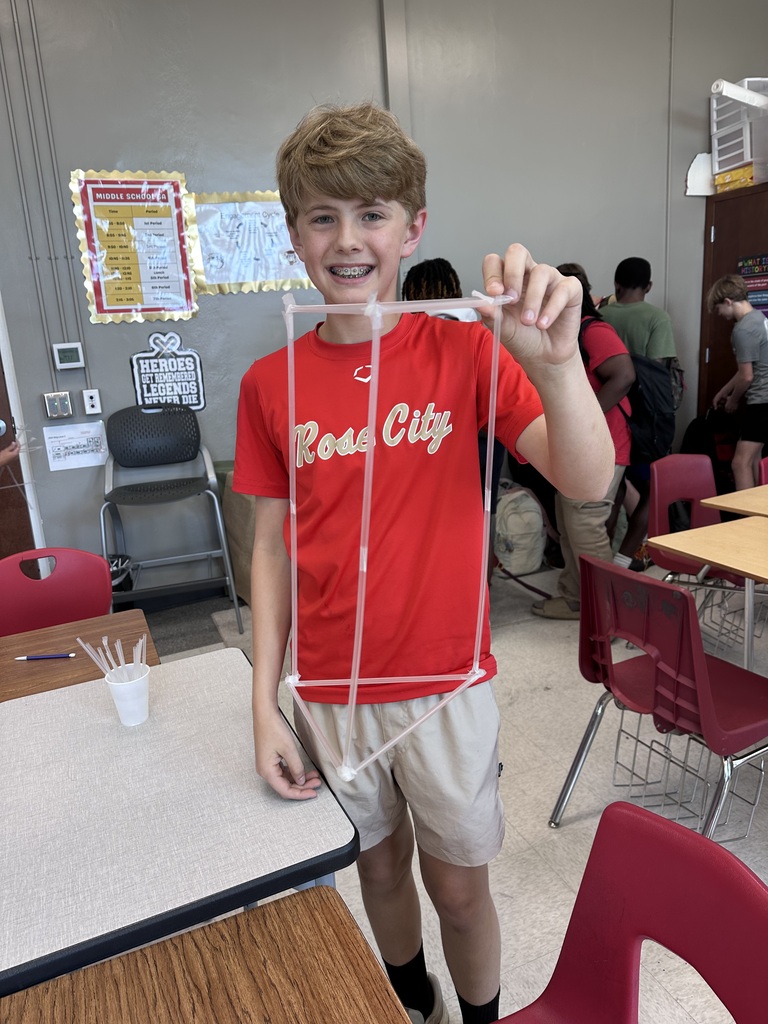 A boy in a red shirt displays a clear model in a classroom with red chairs and tables.