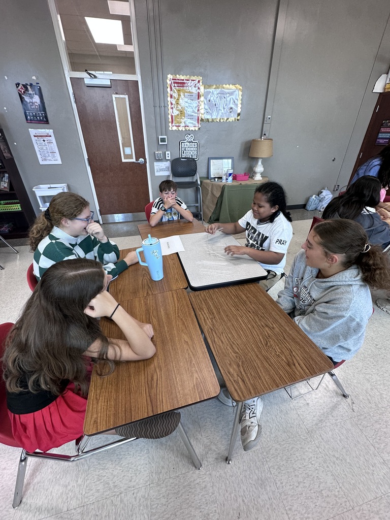 Five young women seated at a table, with papers and a blue cup. A classroom with desks, chairs, and a door.