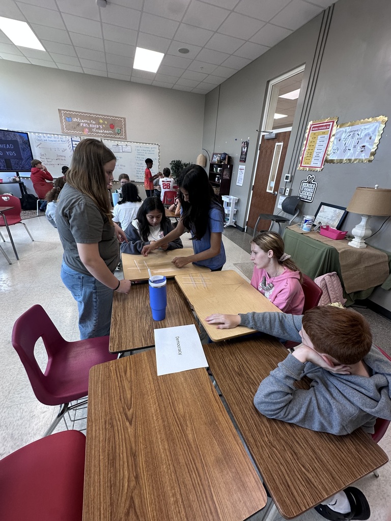 A classroom with students working on a project. Two women stand at a table with materials, while others sit at tables.