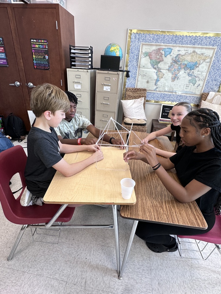 Four students are at a table in a classroom, collaborating on a project. One holds a cup, another has a globe.