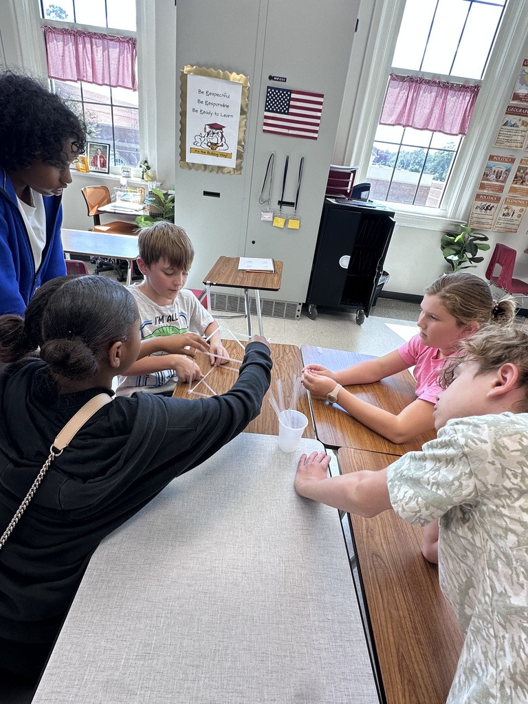 Four children and an adult conduct a science experiment in a classroom with desks, tables, and windows.