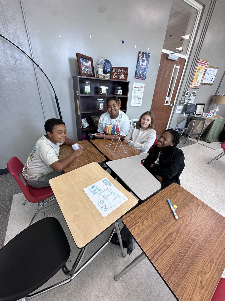 Four students sit around desks in a classroom. Each has a desk and chair. Papers and pencils are on the desks.
