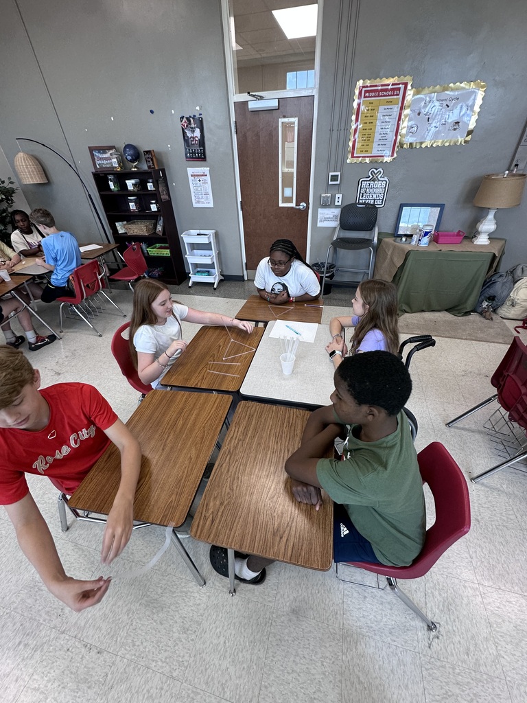 Five students sit at a table in a classroom, looking at papers. Behind them, a teacher sits at a desk.