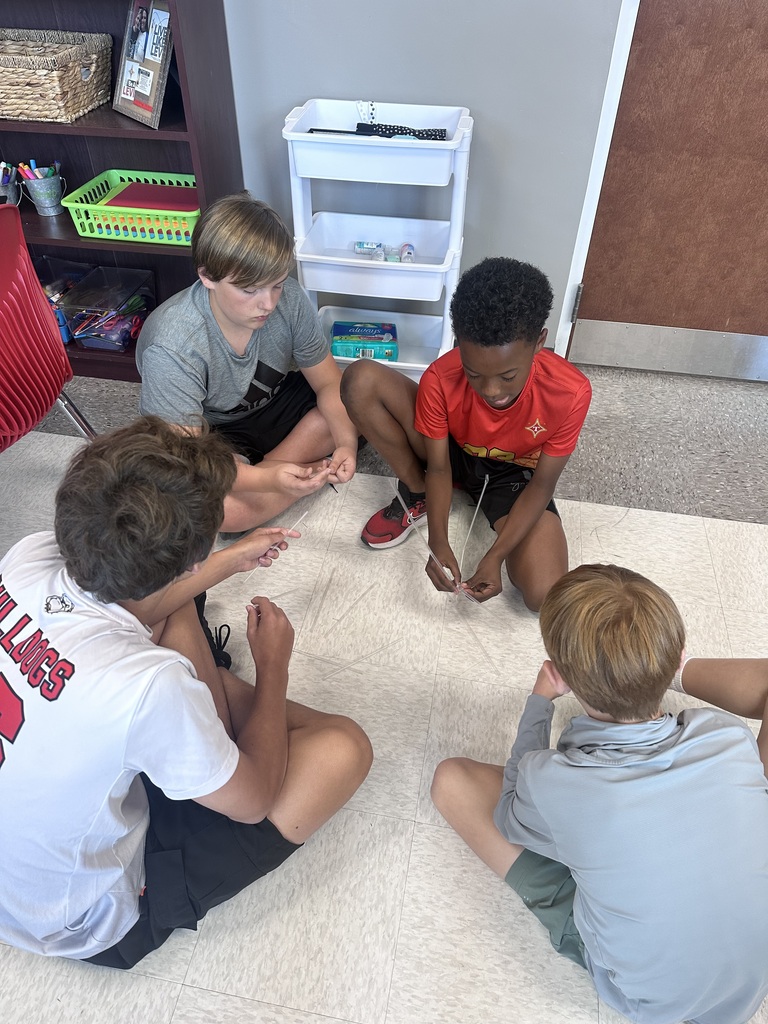 Four boys sit on a white carpet, engaged in an activity. They hold string, and one boy has a red shirt. Behind them, shelves with storage boxes and a red chair are visible.
