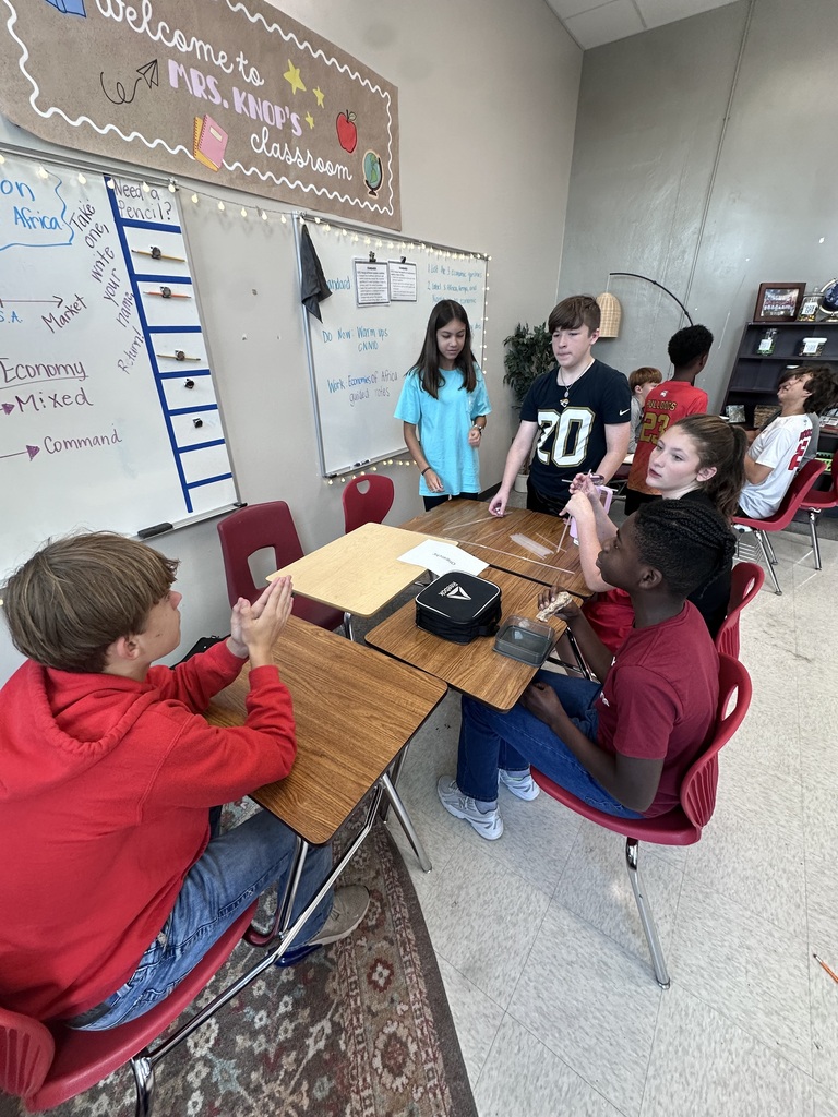 Children in a classroom setting. One student stands by a whiteboard while others sit around a table.