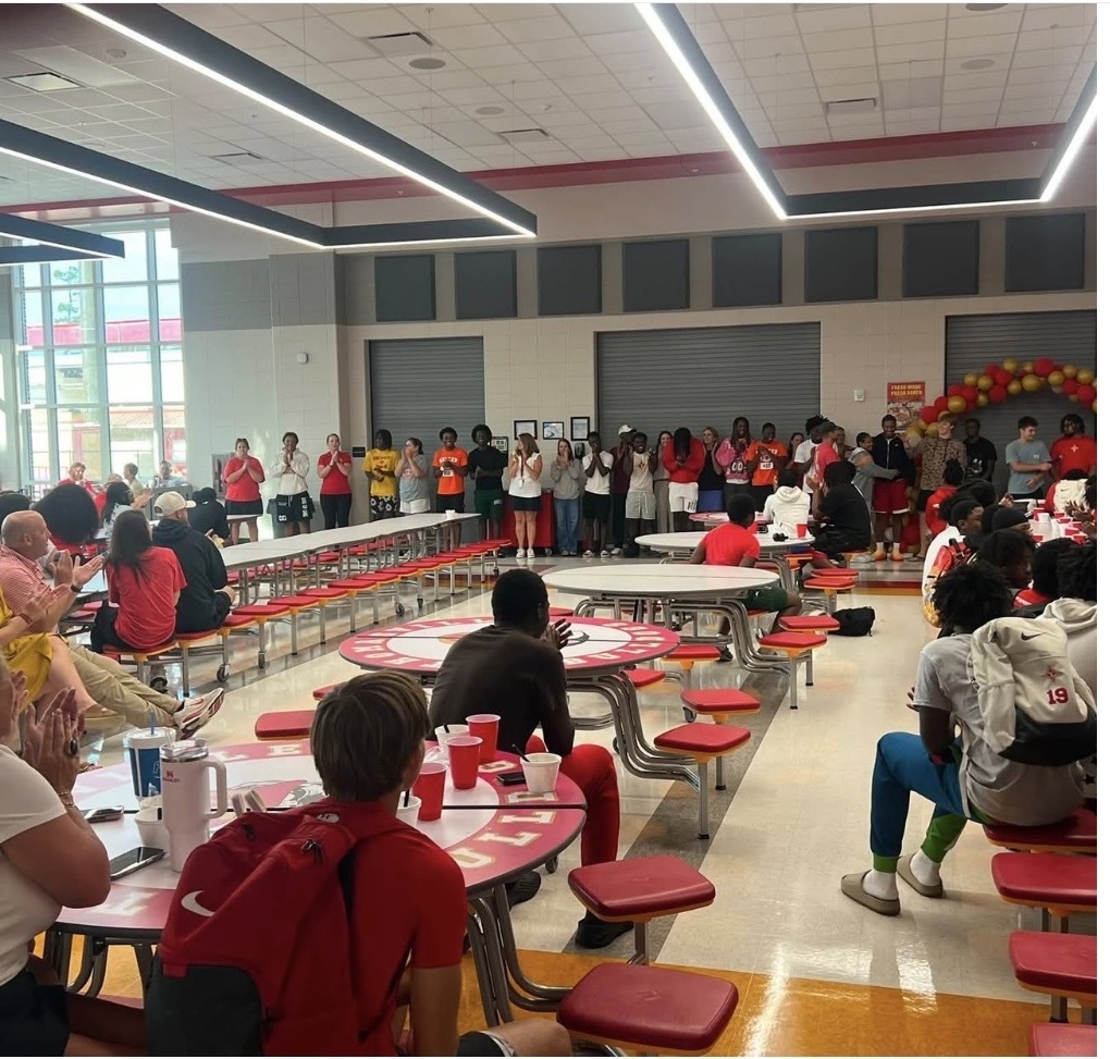 A cafeteria with tables and chairs. People are seated and standing. Some wear backpacks. Red chairs are around.