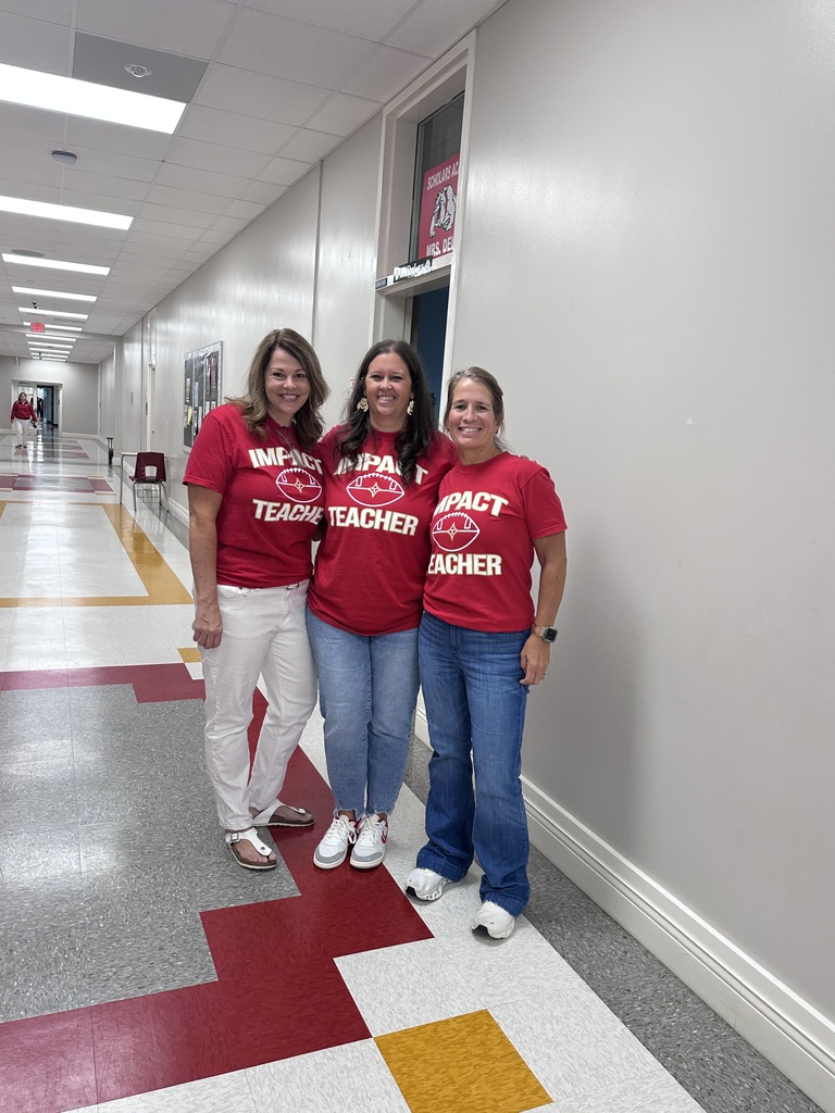 Three women in red shirts stand in a hallway. Two are smiling and looking at the camera.
