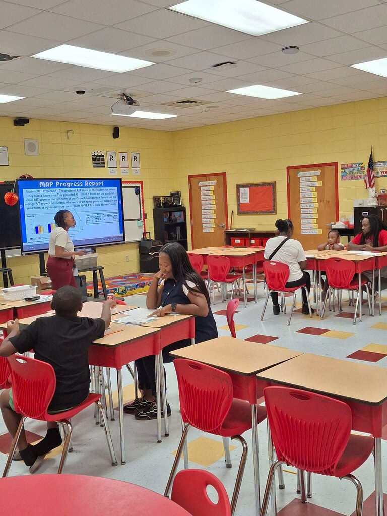Bright classroom with yellow walls and desks. A teacher stands at a projector screen, while students sit at desks, engaged in class.