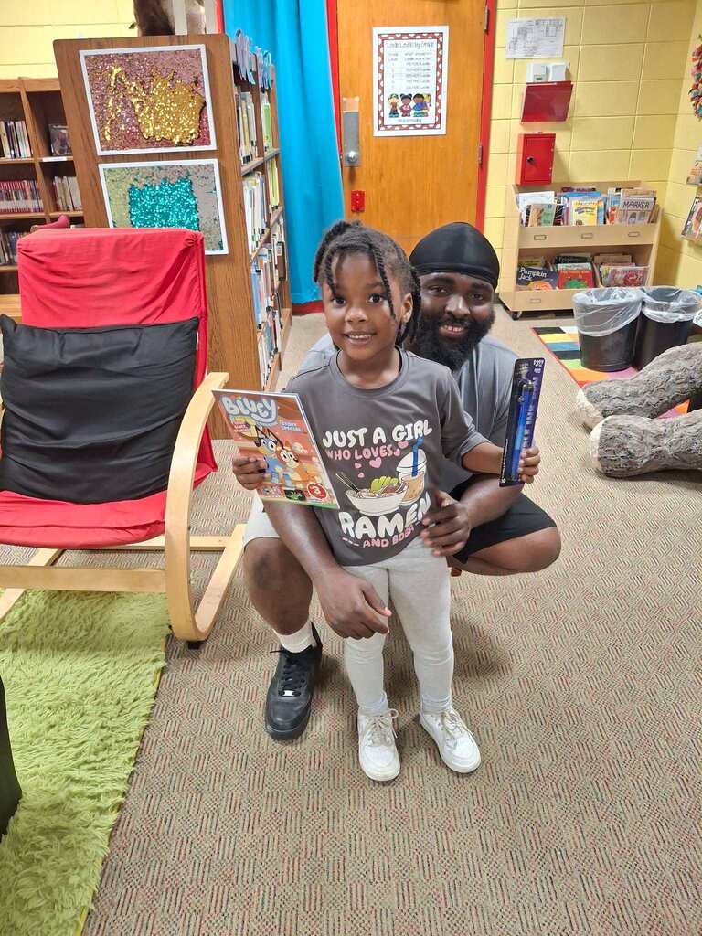 A young child holds a comic book in a library. A person crouches behind the child.