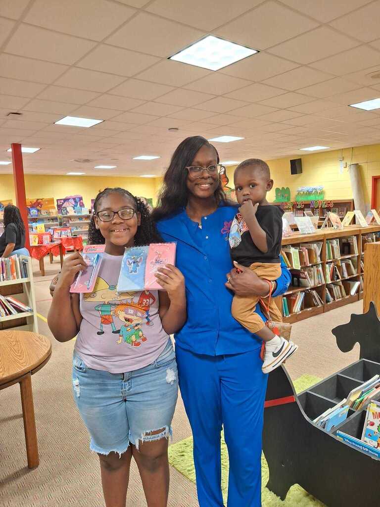 Three people in a library, one holding a book, another holding a baby, and the third holding a book.